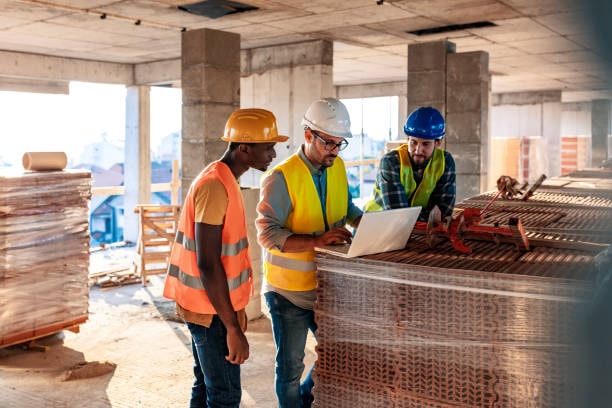 Three construction workers in safety gear reviewing blueprints on commercial building site